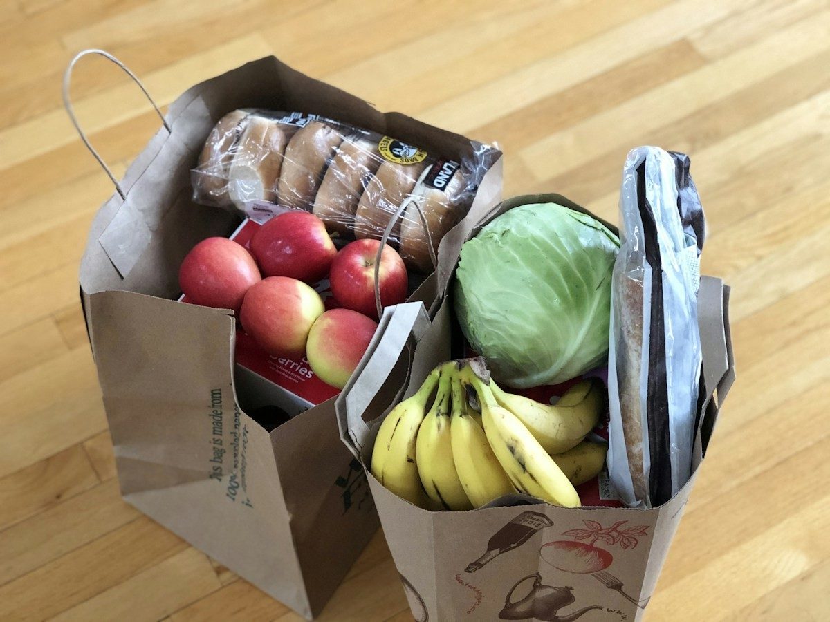 Zwei Papiertüten mit Äpfeln, Bananen, einem Kohlkopf und Brot auf einem Holzboden