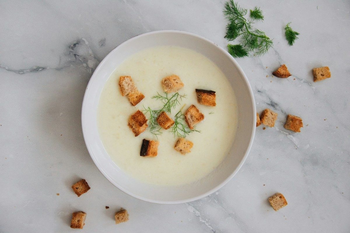 Helle Cremesuppe mit Brotwürfeln und Dill in einer weißen Schale auf Marmor