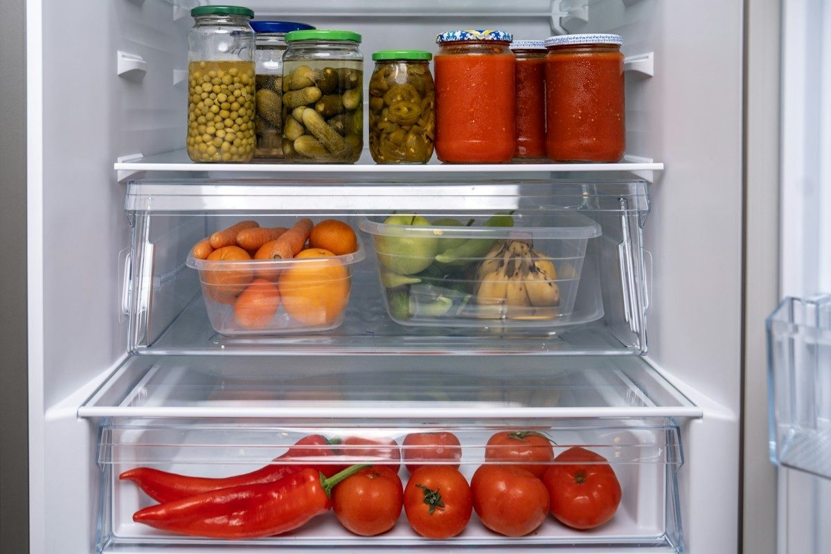 Geöffneter Kühlschrank mit Gläsern oben, Obst in der Mitte und Tomaten und Paprika im Gemüsefach