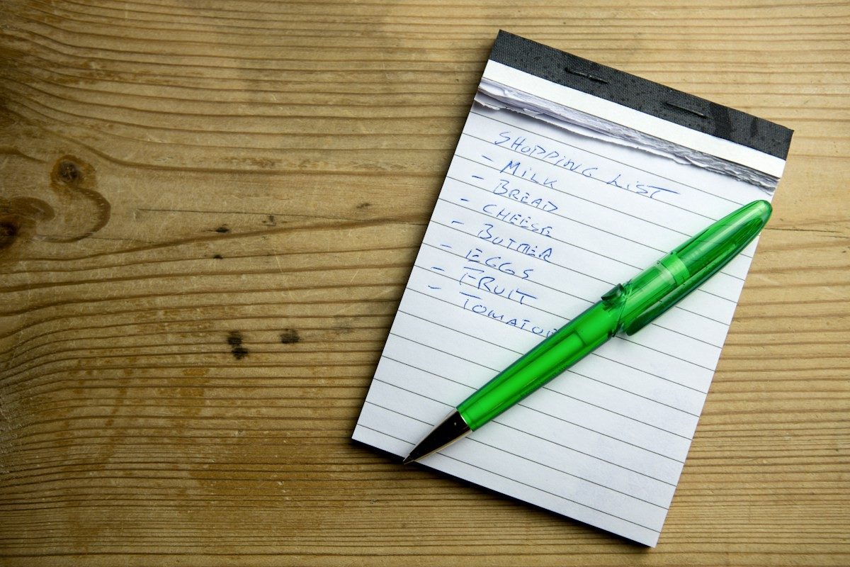 Notizblock mit einer handgeschriebenen Einkaufsliste und einem grünen Stift auf Holz